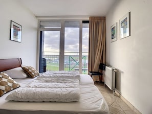 A hotel room with a bed, a radiator, a chair, and a view of a green landscape through large windows.