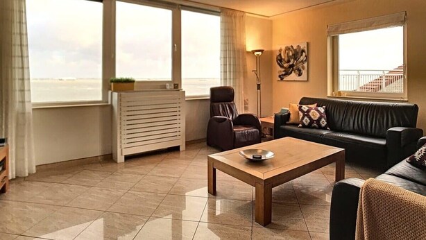 Een ruime woonkamer met een groot raam, een bank, een fauteuil, een salontafel en een radiator.