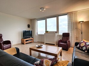 Una sala de estar moderna con un sofá, dos sillones, una mesa de centro, un televisor y un amplio ventanal con cortinas.