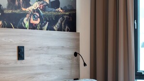 Moderni hotellihuone, jossa on suuri sänky, vaaleasta puusta valmistettu pääty, musta seinävalaisin ja ikkuna, jossa on beigenväriset verhot.