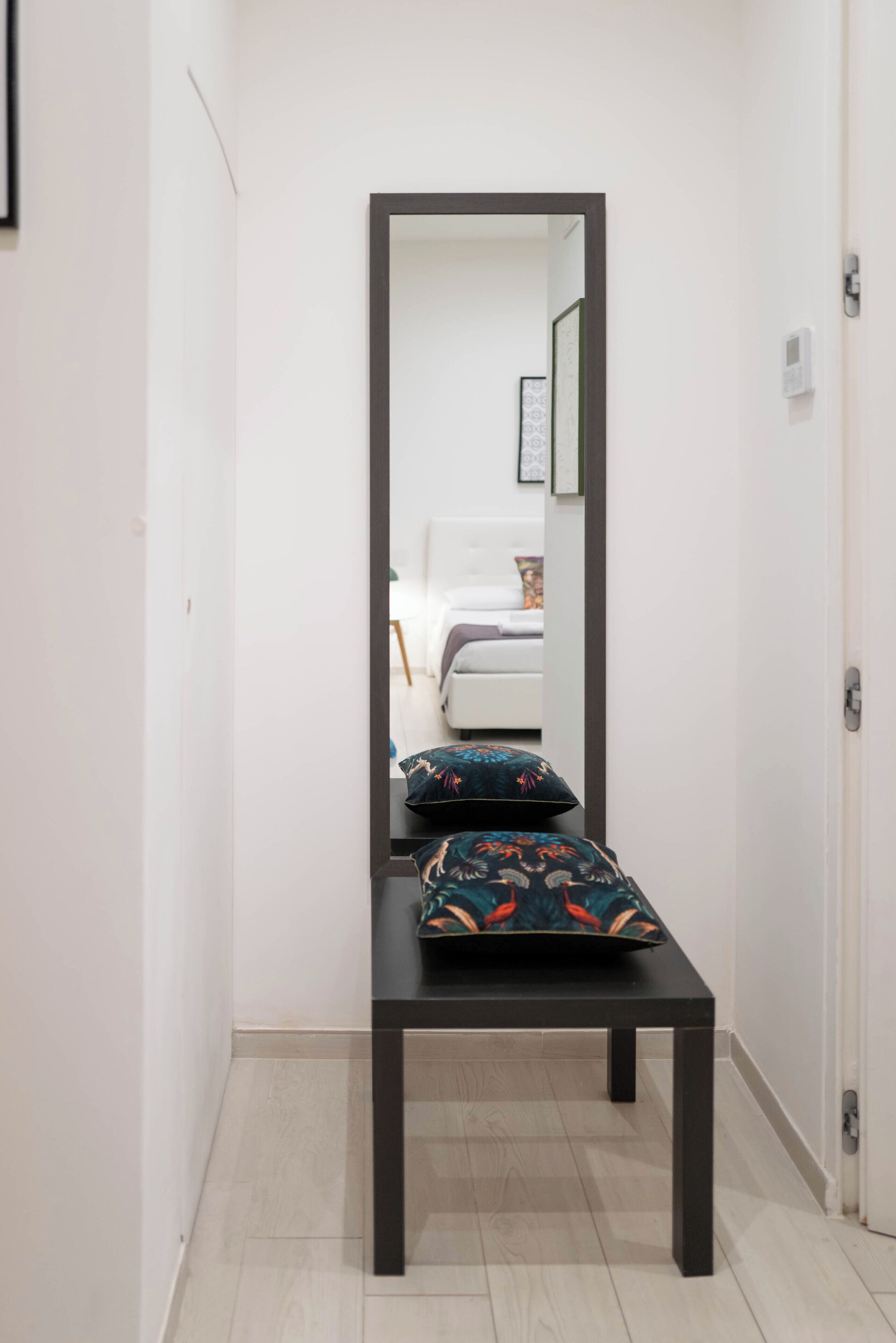 A narrow hallway with a bench and a mirror reflecting a bed and a bedside table.