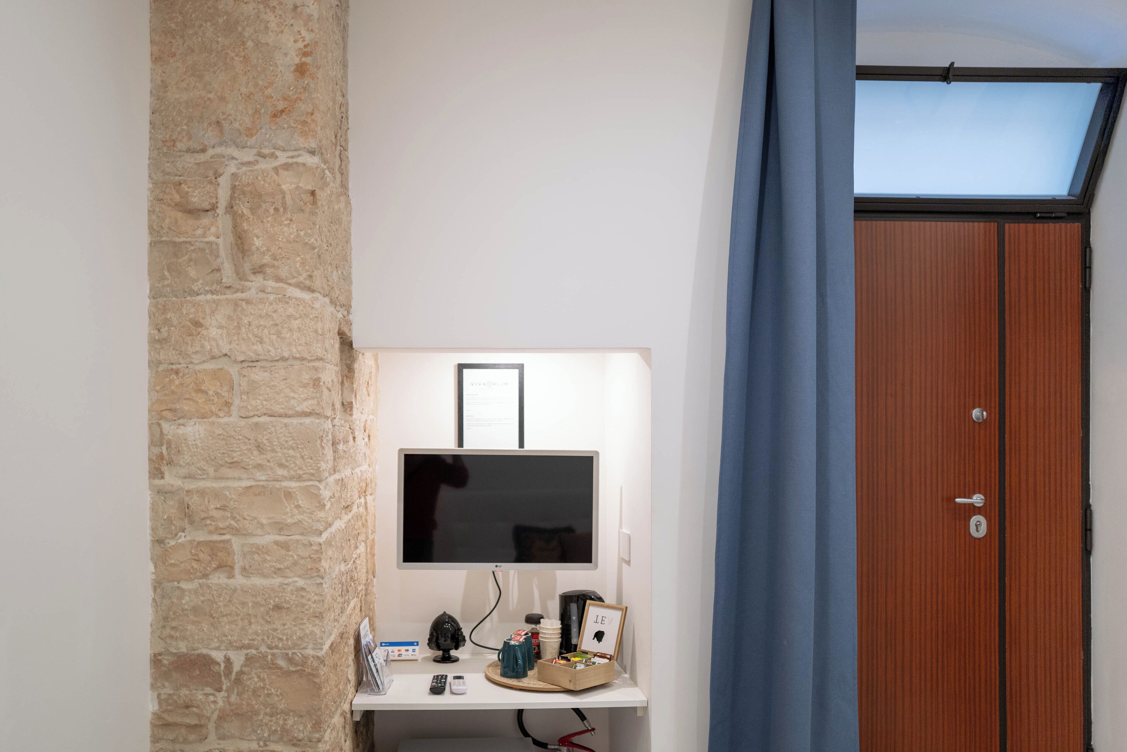 Una habitación con pared de piedra, puerta de madera, un escritorio blanco con monitor de computadora, un control remoto y un estante con objetos.