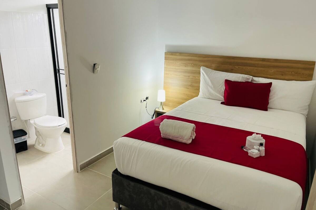 A hotel room with a bed, a nightstand, a lamp, a red pillow, and a white toilet.