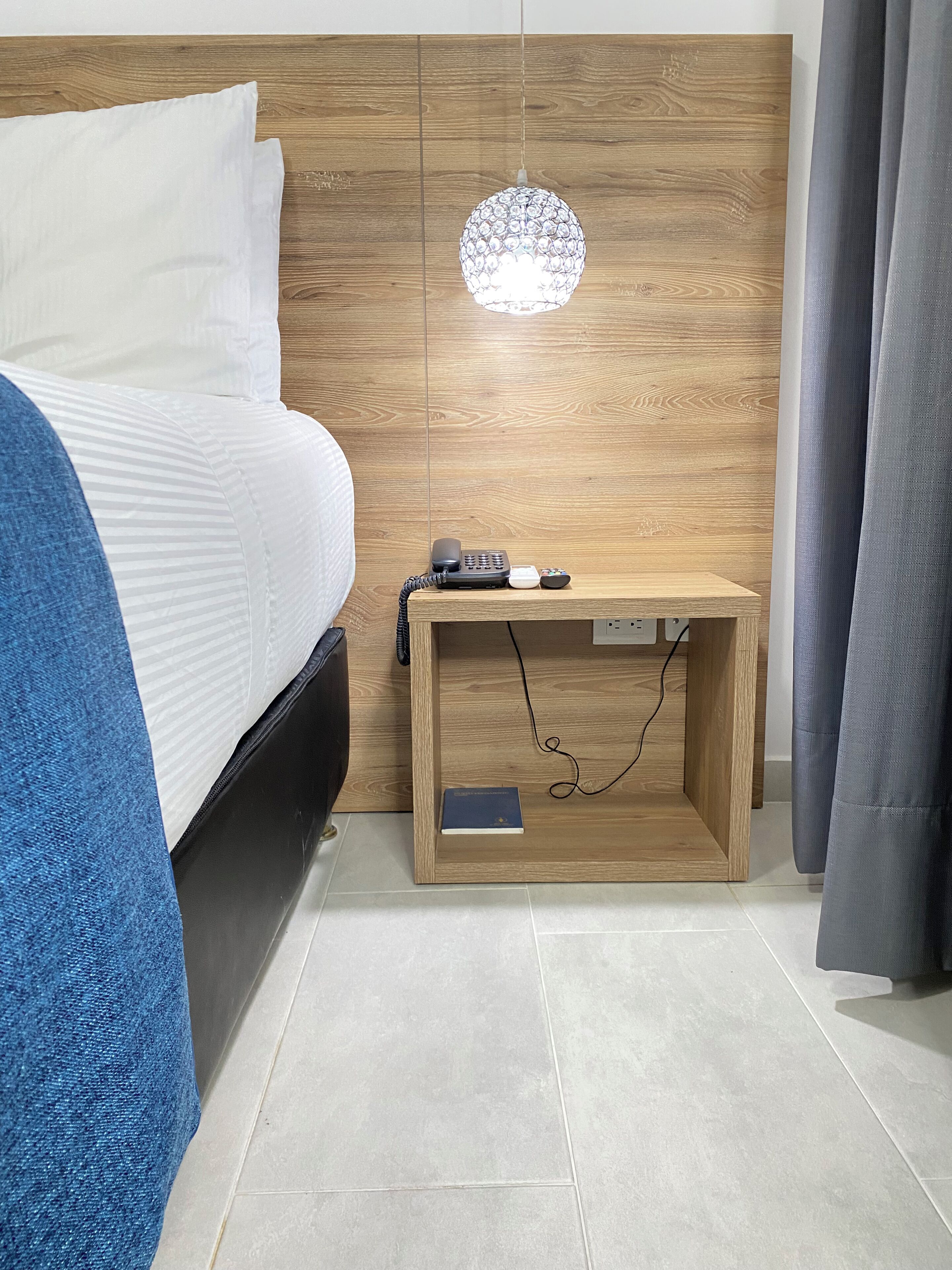 Una cama bien tendida con sábanas blancas, cabecera de madera, mesita de noche con teléfono y control remoto, y una lámpara colgante.