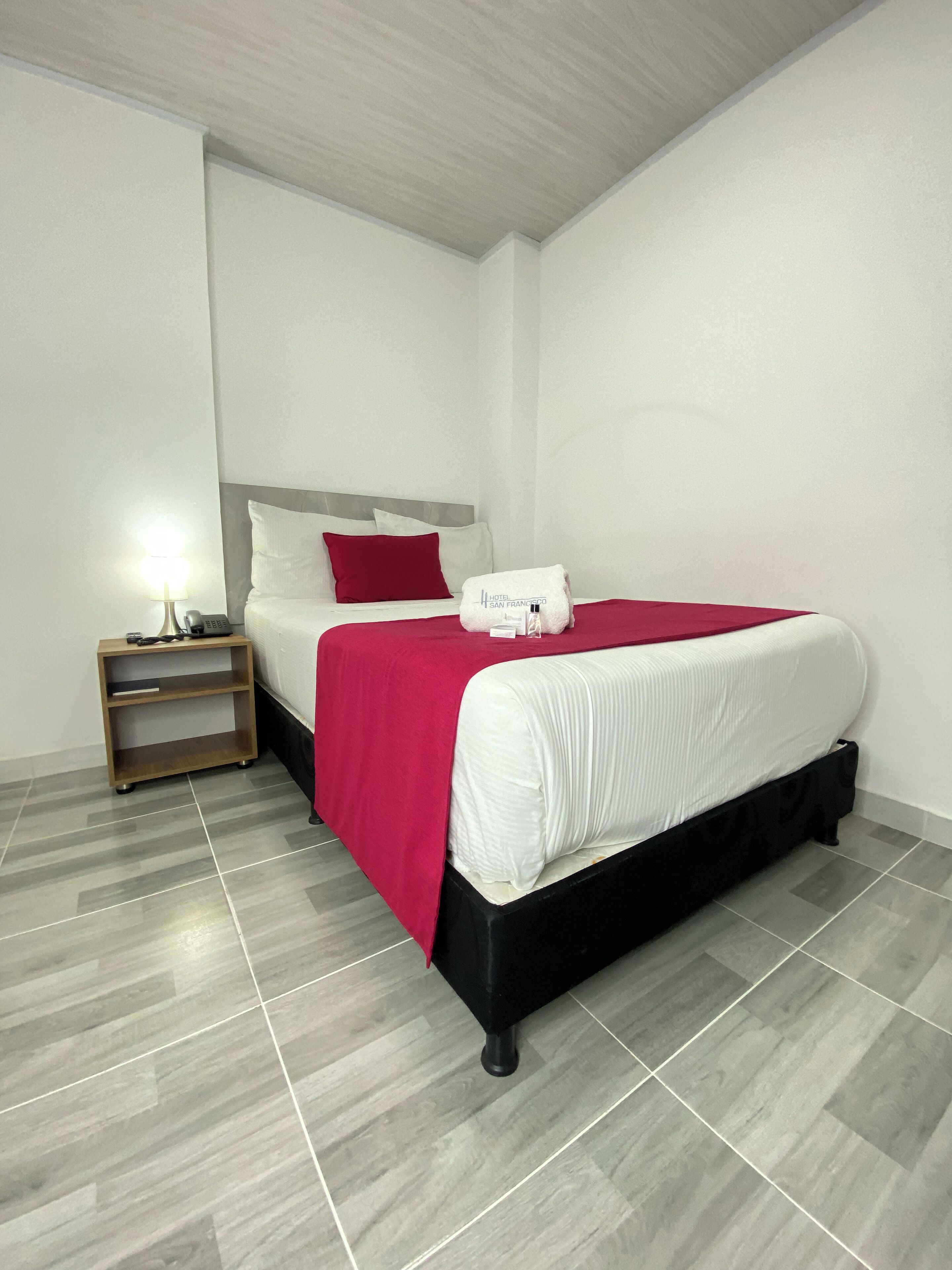 A hotel room with a bed, a nightstand with a lamp, and a red pillow.