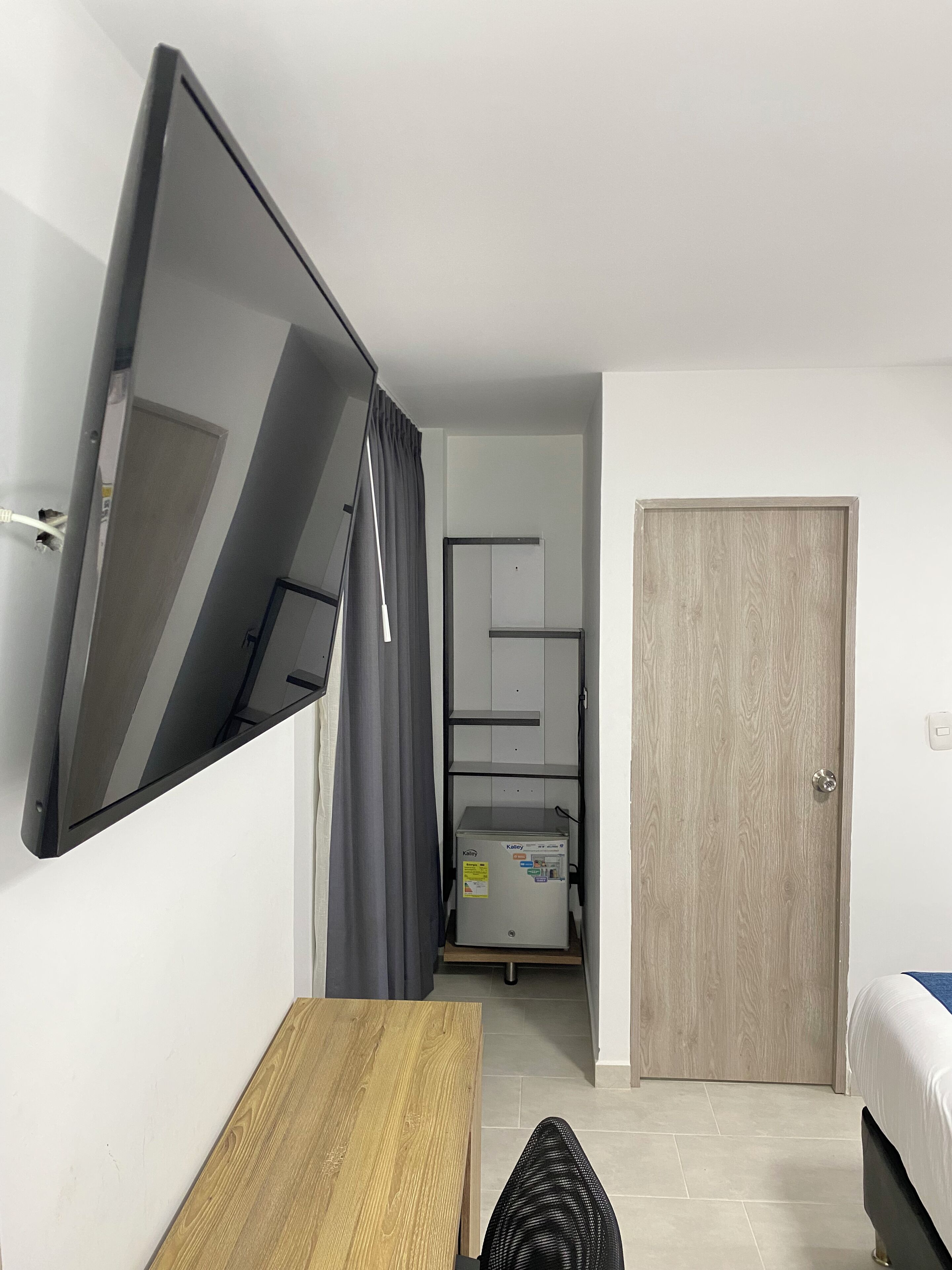 A compact hotel room with a flat-screen TV mounted on the wall, a wooden desk, a bed with a white comforter, and a wooden door.