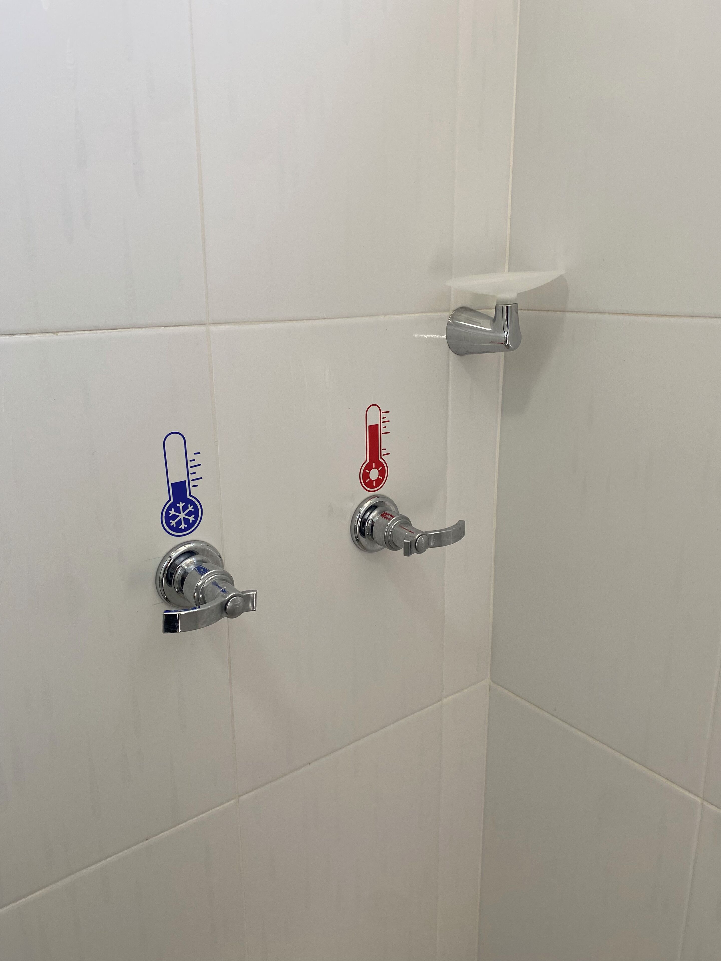 A bathroom with white tiled walls, a shower area, and two faucets with temperature control icons.