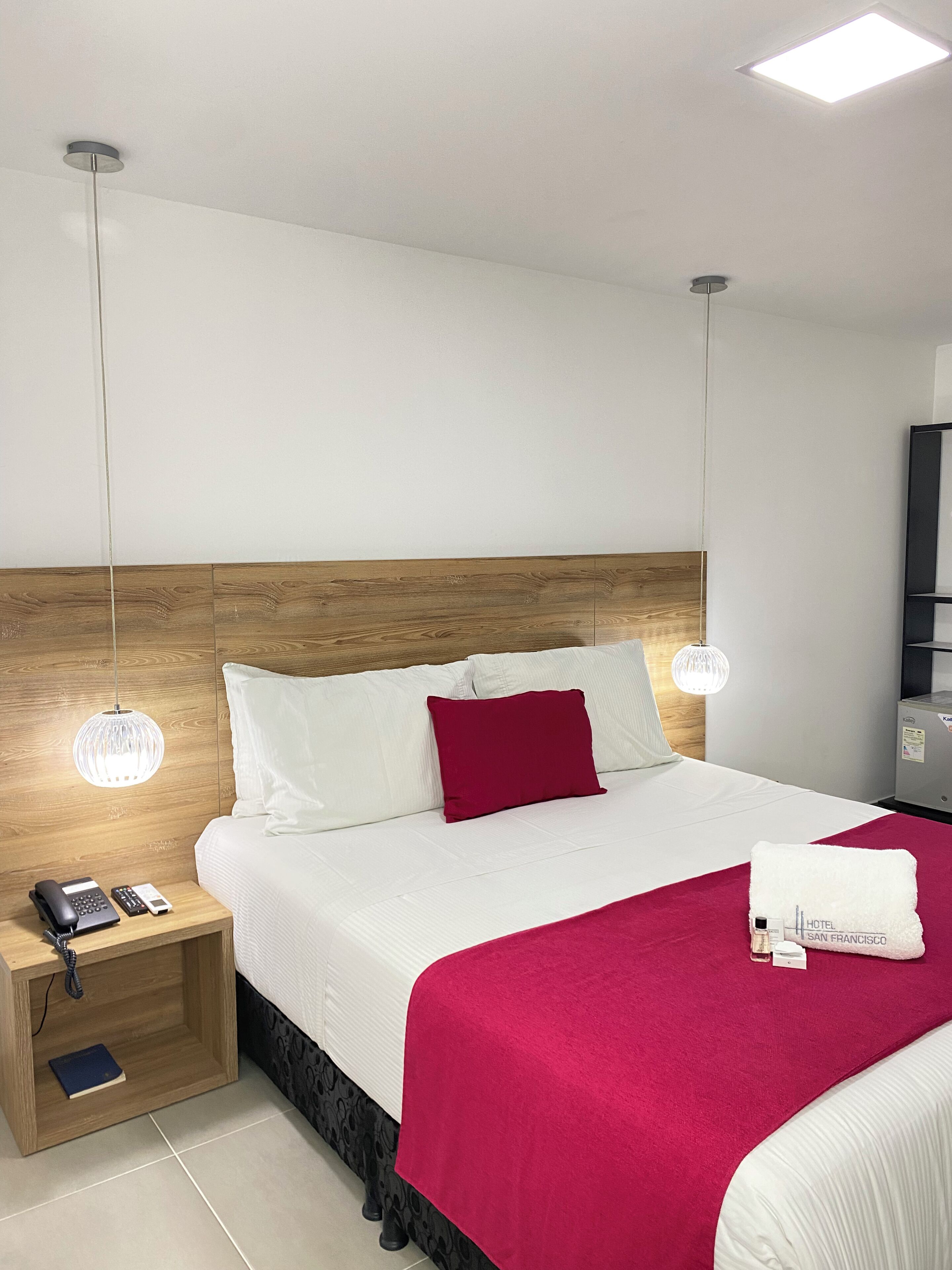A hotel room with a wooden headboard, white bedding, a red throw blanket, a white towel with a hotel name tag, a bedside table with a phone and a book, and a hanging light fixture.