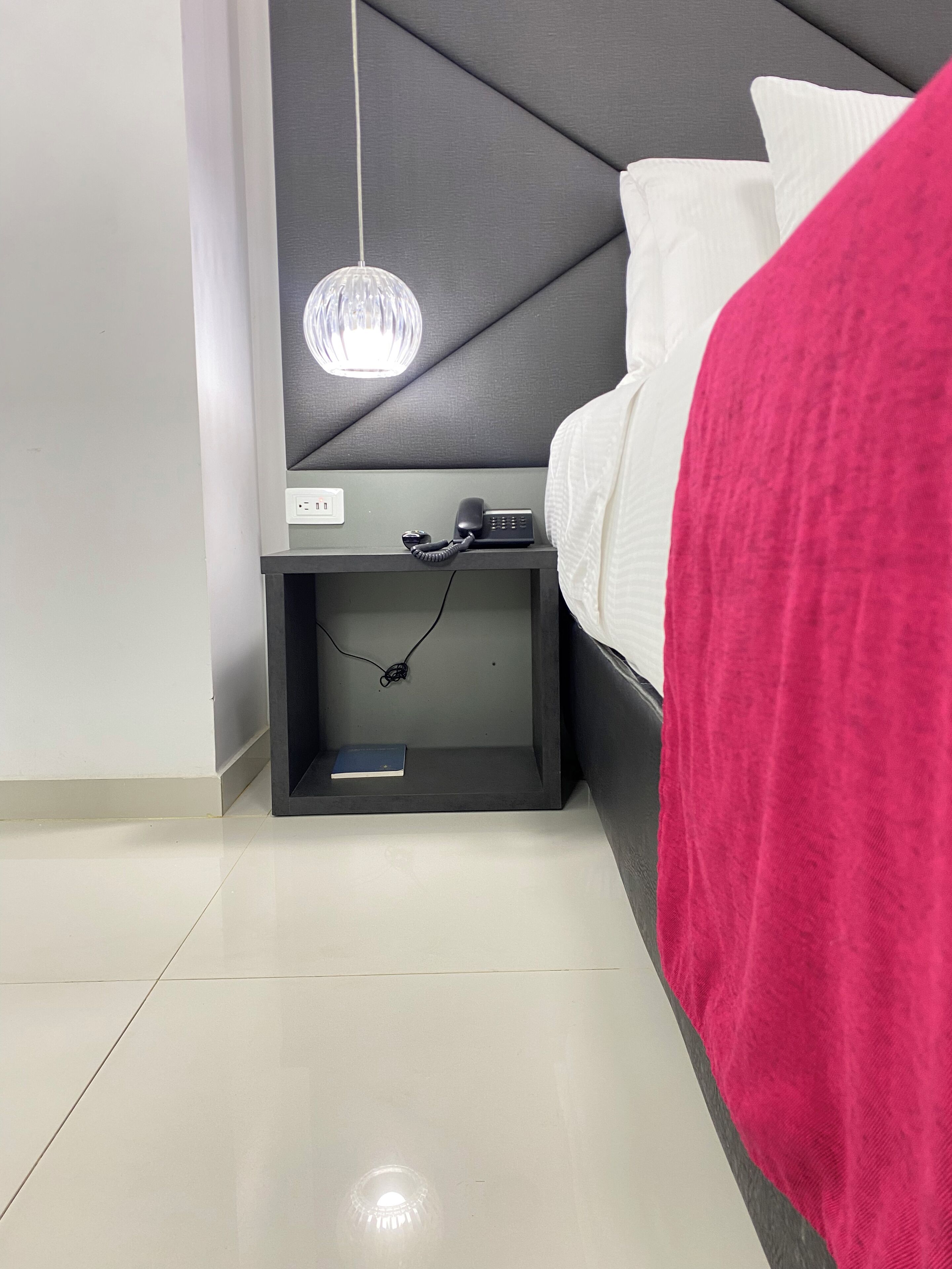 A modern hotel room with a grey headboard, a bedside table with a phone and a book, and a hanging light fixture.