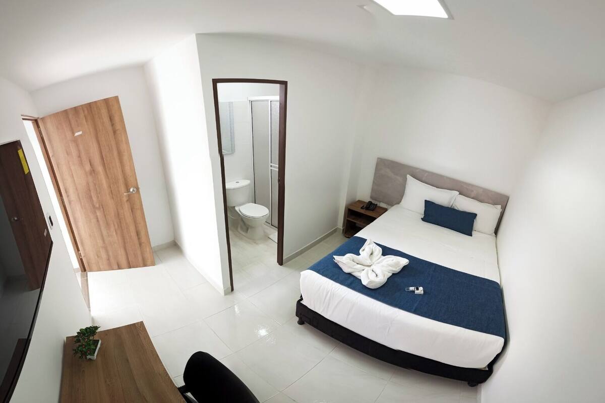 A modern hotel room with a large bed, a wooden door, a small bathroom, and a desk with a chair.
