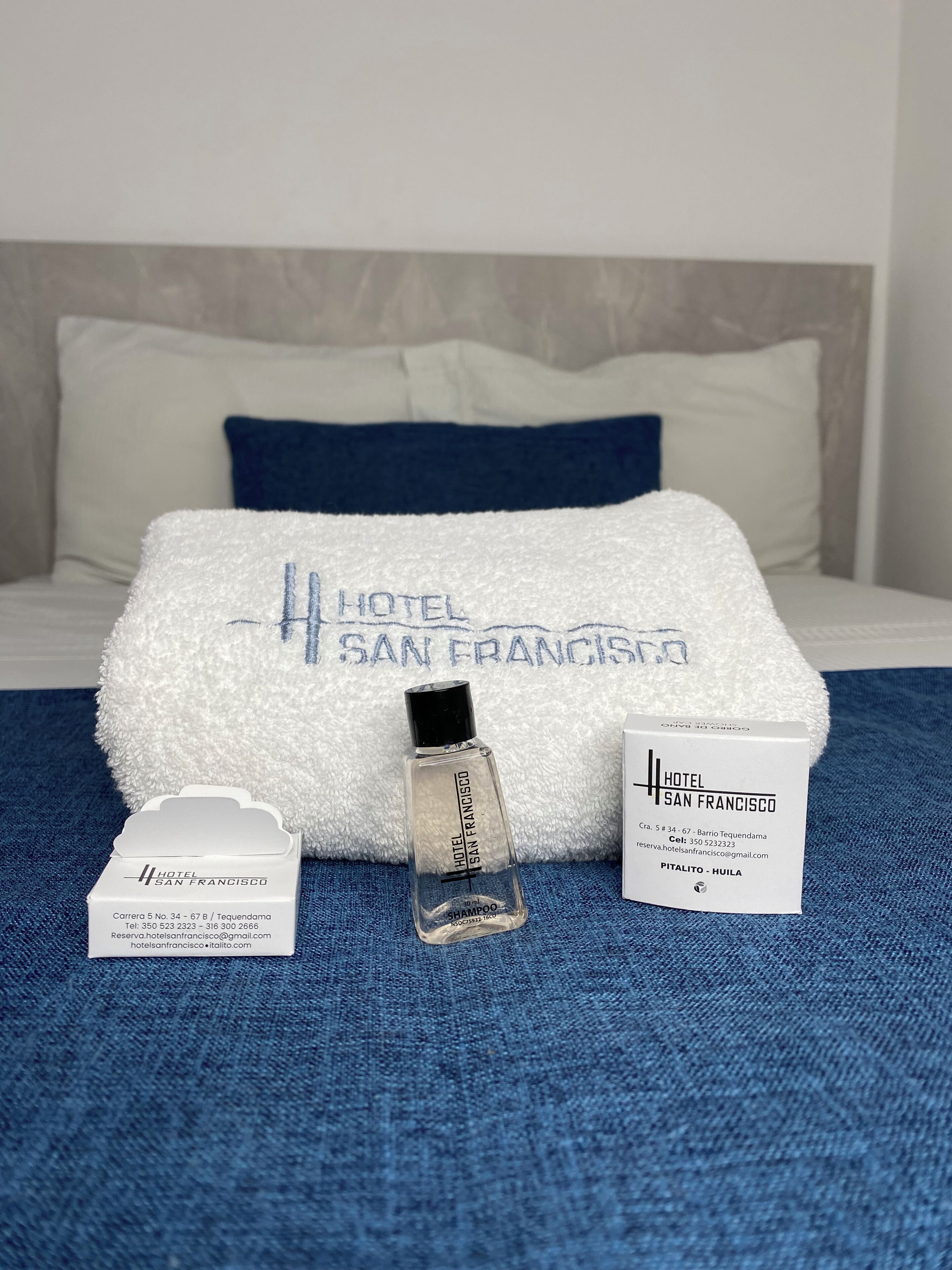 A neatly made bed with a white towel and a clear bottle of shampoo, a white hotel information card, and a blue pillow.