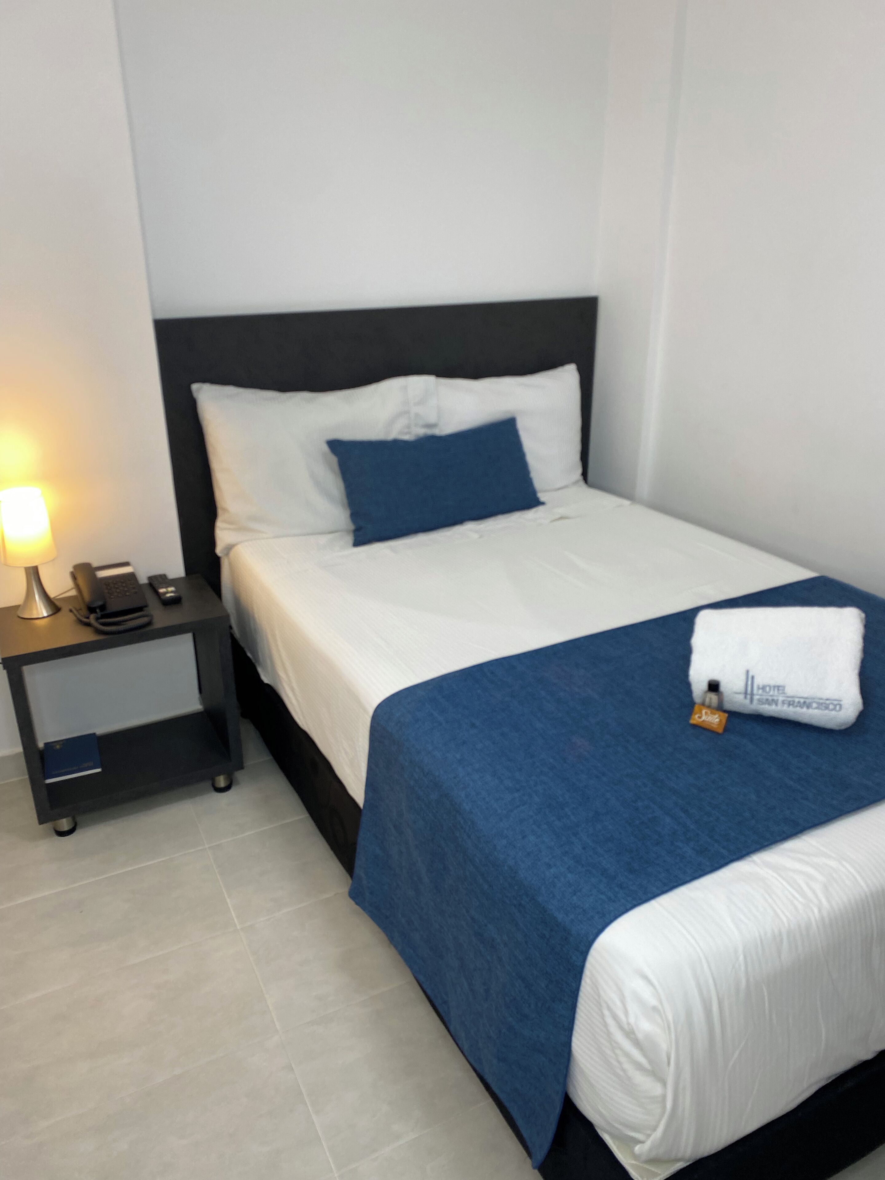 Un lit bien fait, avec une literie blanche et bleue, une lampe de chevet, une petite table de nuit avec un téléphone et un livre, et une serviette pliée avec une étiquette d’hôtel.