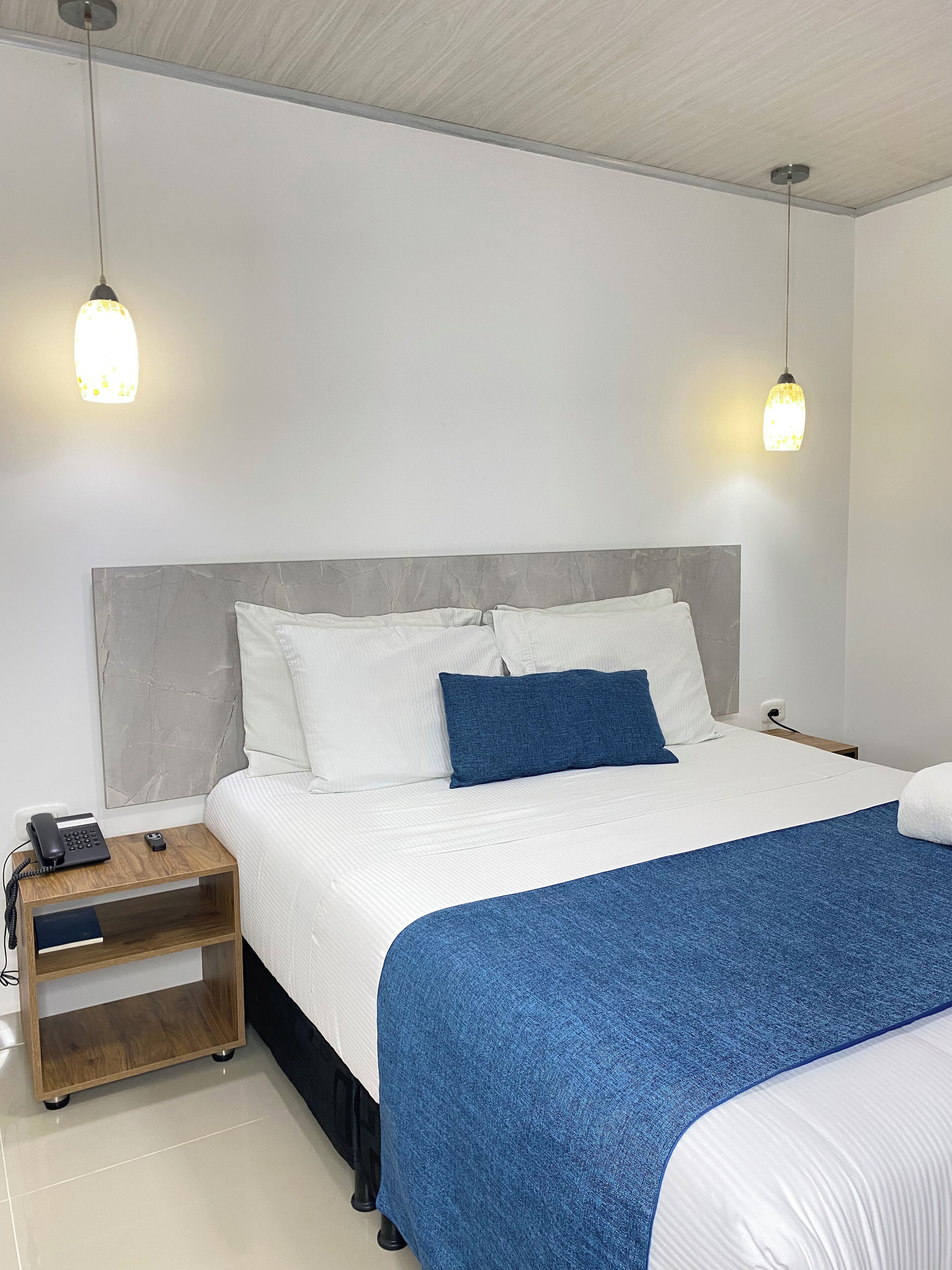 Una habitación de hotel ordenada con una cama, mesitas de noche y dos lámparas colgantes.
