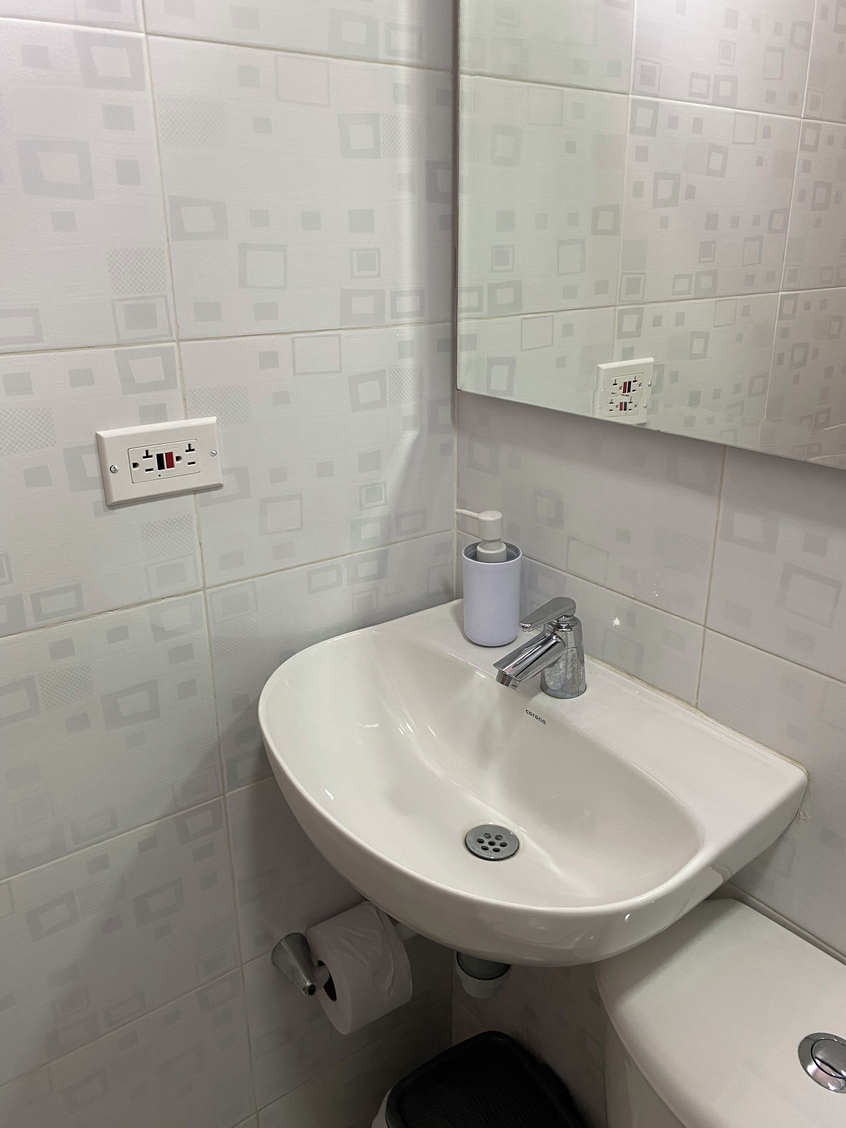 Een badkamer met een wit wastafel, een spiegel en een toilet. Er ligt een rol toiletpapier en staat een prullenbak.