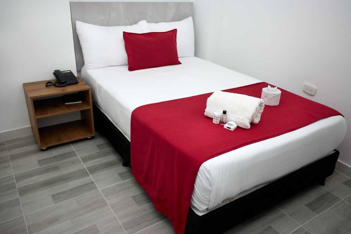 A hotel room with a bed, a nightstand, a red throw blanket, and a white towel.