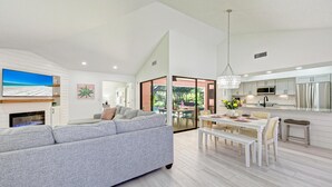 Una sala moderna con un sofá gris, un comedor con mesa blanca y una cocina con electrodomésticos de acero inoxidable.