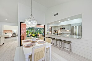 Une cuisine moderne dotée d’appareils électroménagers en acier inoxydable, un coin repas avec une table blanche dressée, et une vue sur un jardin grâce à de grandes fenêtres.