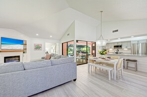 Ein modernes Wohnzimmer mit einem grauen Sofa, einem Essbereich mit einem weißen Tisch und einer Küche mit weißen Schränken und Edelstahlgeräten.