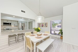 A modern kitchen with stainless steel appliances, a dining area with a white table and bench, and a large window with blinds. - Heated Pool + Game Room + Free EV Charging near Anna Maria Island & IMG Academy (Bradenton)