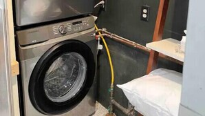 A compact laundry area with two stacked front-loading washing machines, a shelf with a detergent box, and a visible water pipe. - Newly Renovated Downtown Gem! (Buffalo)