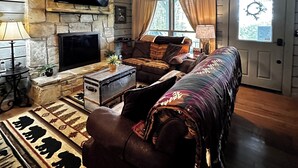 Un salon chaleureux avec une cheminée en pierre, un téléviseur à écran plat, un ventilateur de plafond et une grande fenêtre.