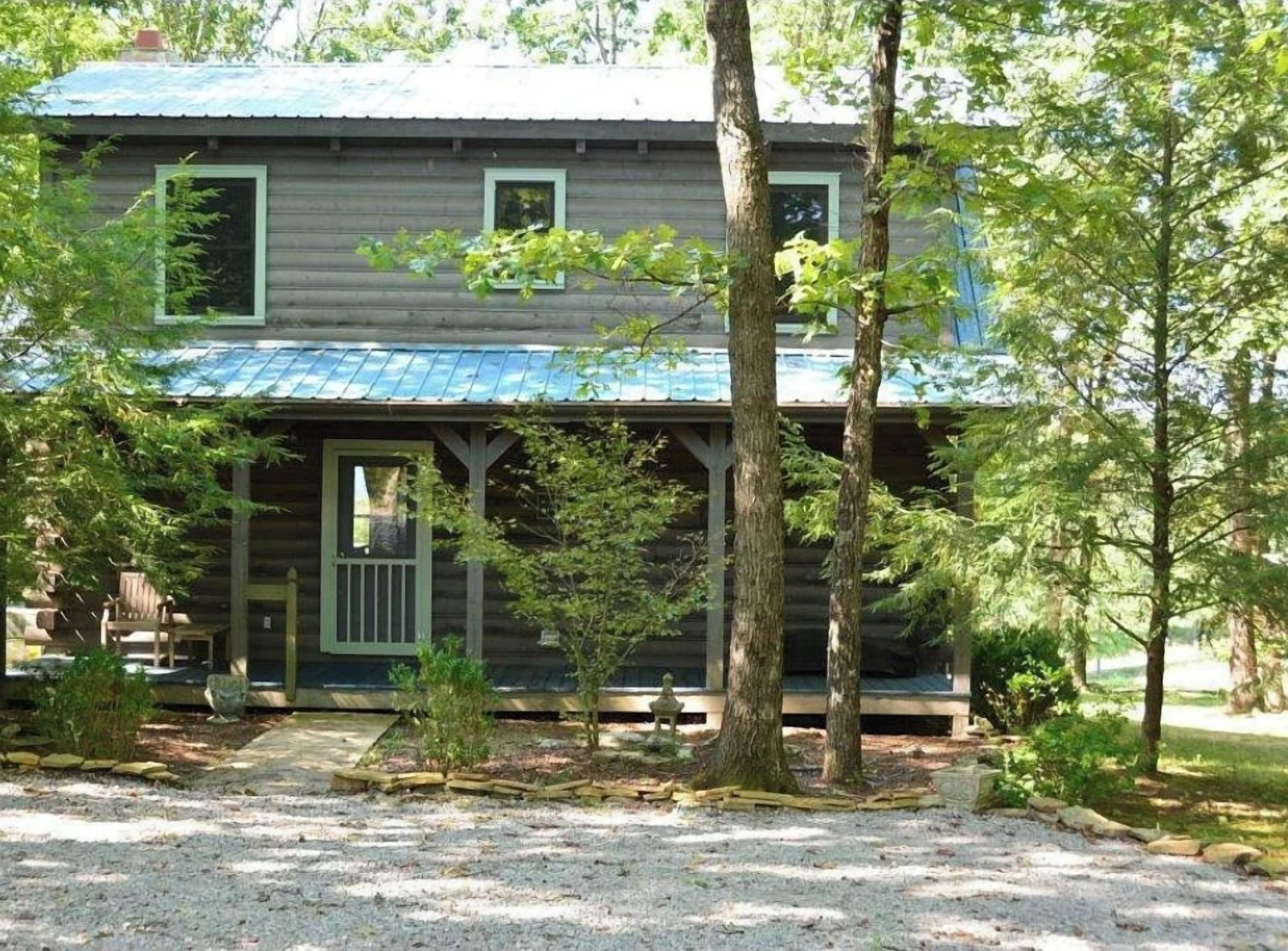 Une cabane rustique en rondins de bois, dotée d’un porche, entourée d’arbres et bordée d’une allée en gravier.