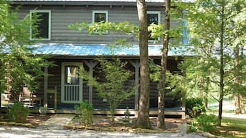 Une cabane rustique en rondins de bois, dotée d’un porche, entourée d’arbres et bordée d’une allée en gravier.