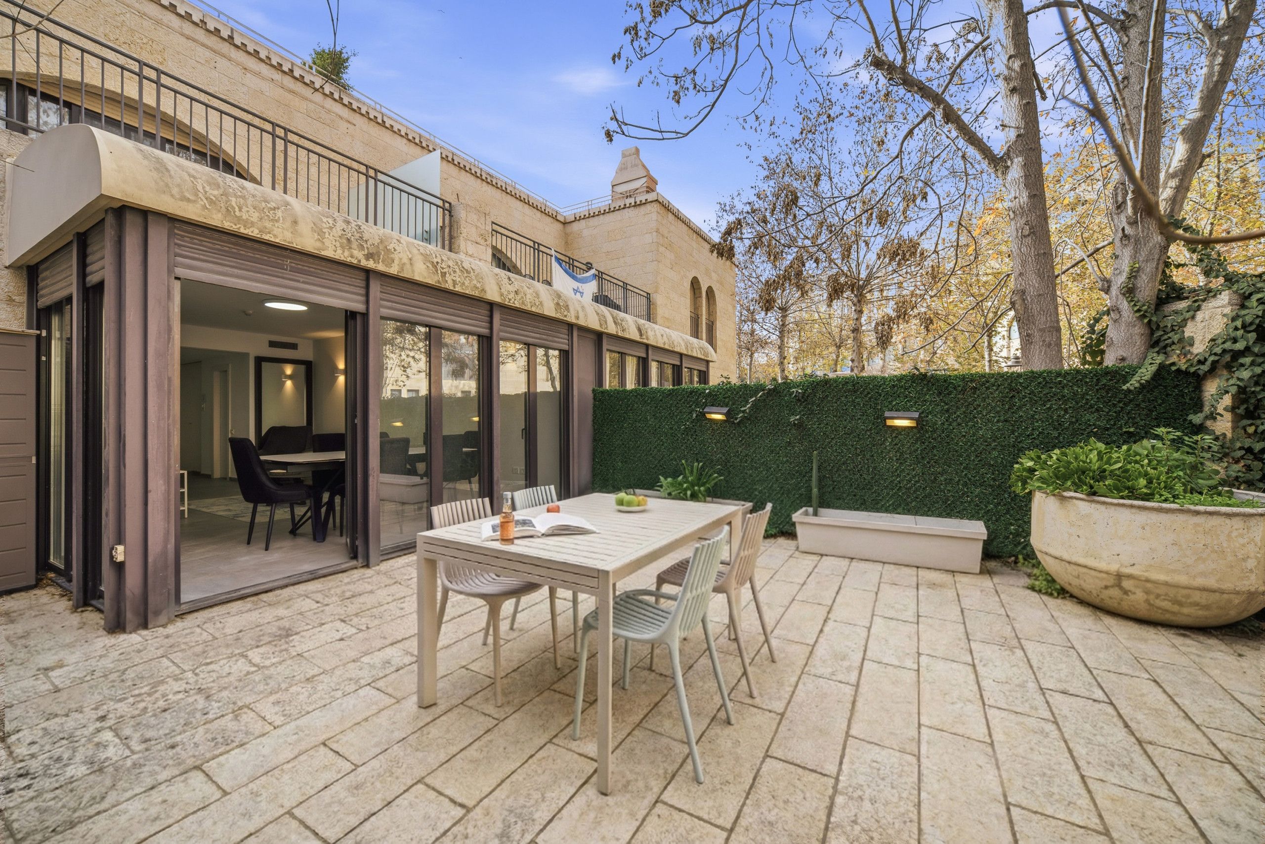 A patio with a table and chairs, a green hedge, and a building with a balcony.