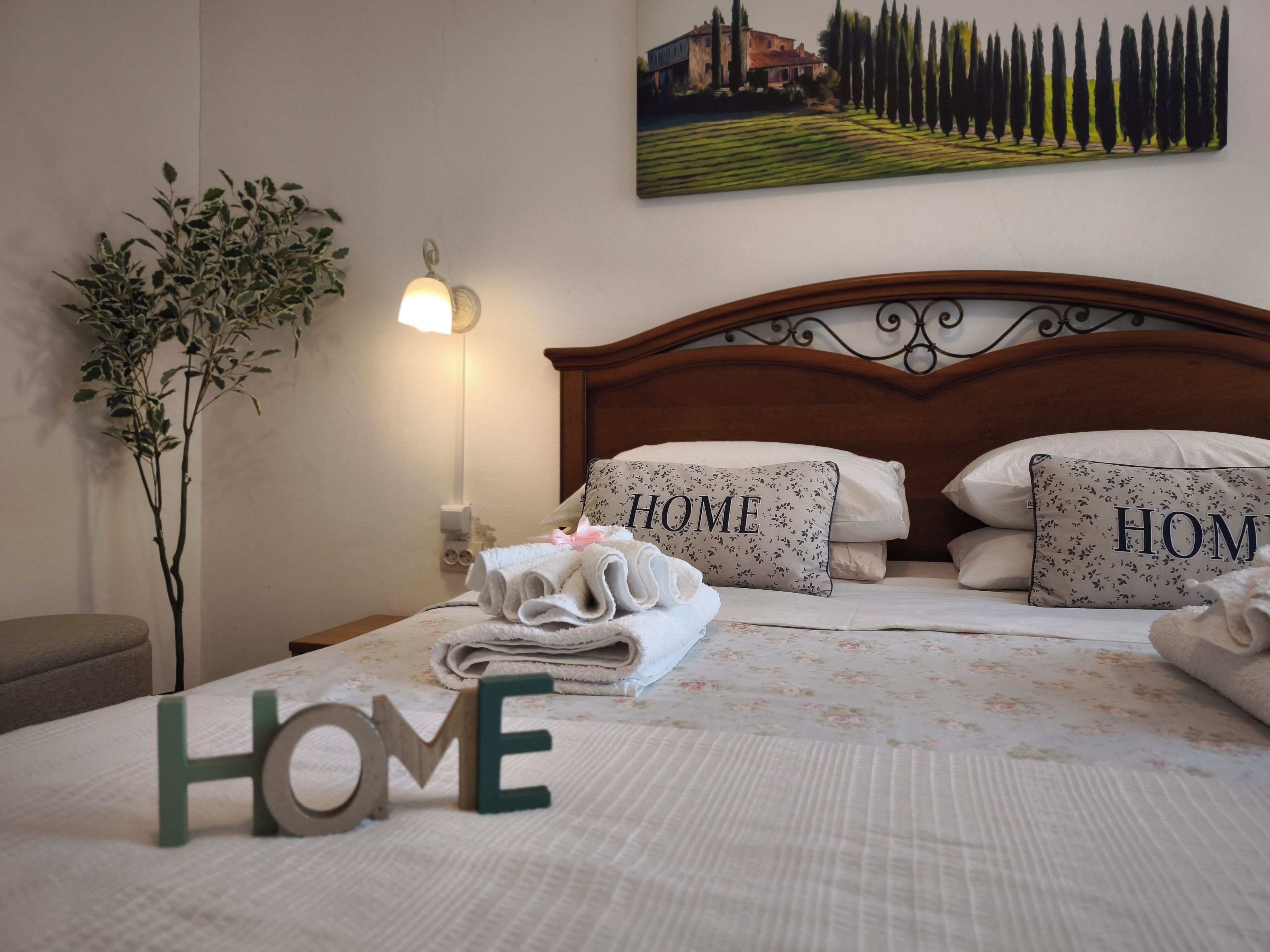 Una cama bien hecha con almohadones que dicen 'HOME' y un cartel con la palabra 'HOME', un cuadro colgado en la pared, una lámpara de noche y una planta en maceta.