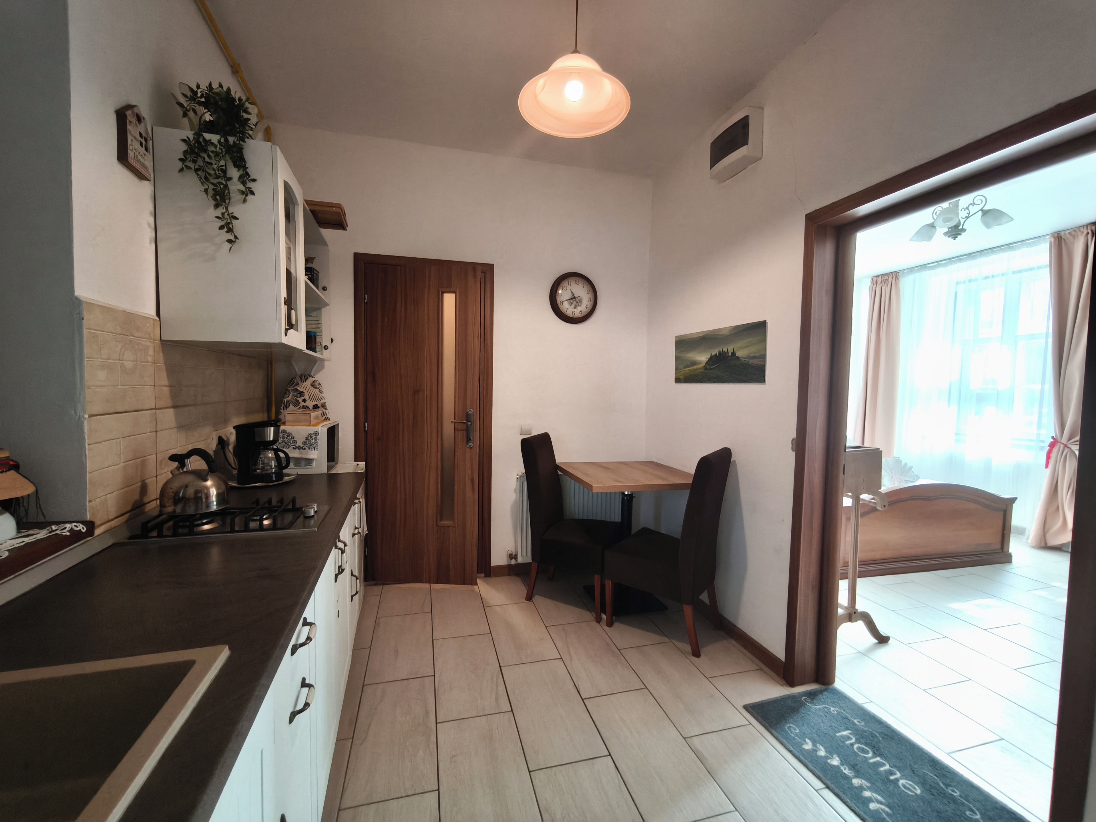 A kitchen with a wooden door, a dining table with chairs, a clock on the wall, and a view through a window.
