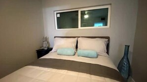A bedroom with a bed, two pillows, a nightstand, a vase, and a window above the bed. - Little Blue Box  (Phoenix)