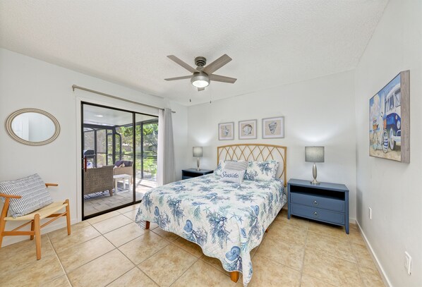 Quarto com cama, cadeira, mesa de cabeceira, ventoinha no teto e vista por uma porta de vidro.