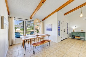 Uma área de refeições com mesa de madeira e bancos, luminária pendente e vista através de grandes janelas.
