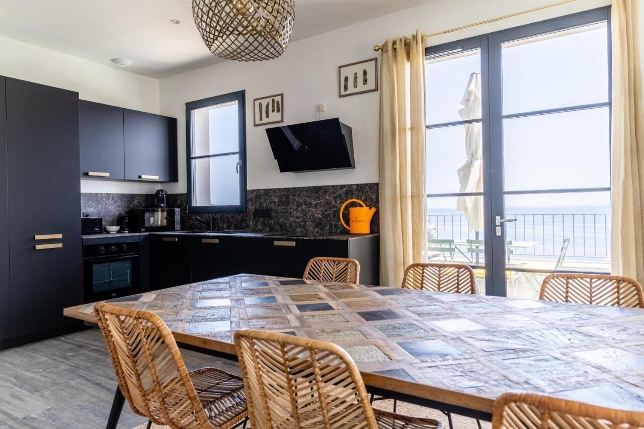 Une cuisine moderne avec des meubles de couleur sombre, un comptoir en marbre, une table à manger recouverte d’une nappe à carreaux, des chaises en osier et une grande fenêtre offrant une belle vue.