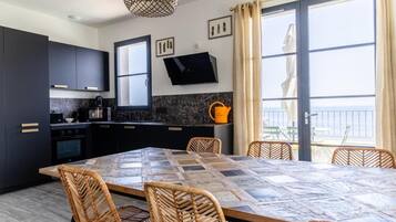 Une cuisine moderne avec des meubles de couleur sombre, un comptoir en marbre, une table à manger recouverte d’une nappe à carreaux, des chaises en osier et une grande fenêtre offrant une belle vue.