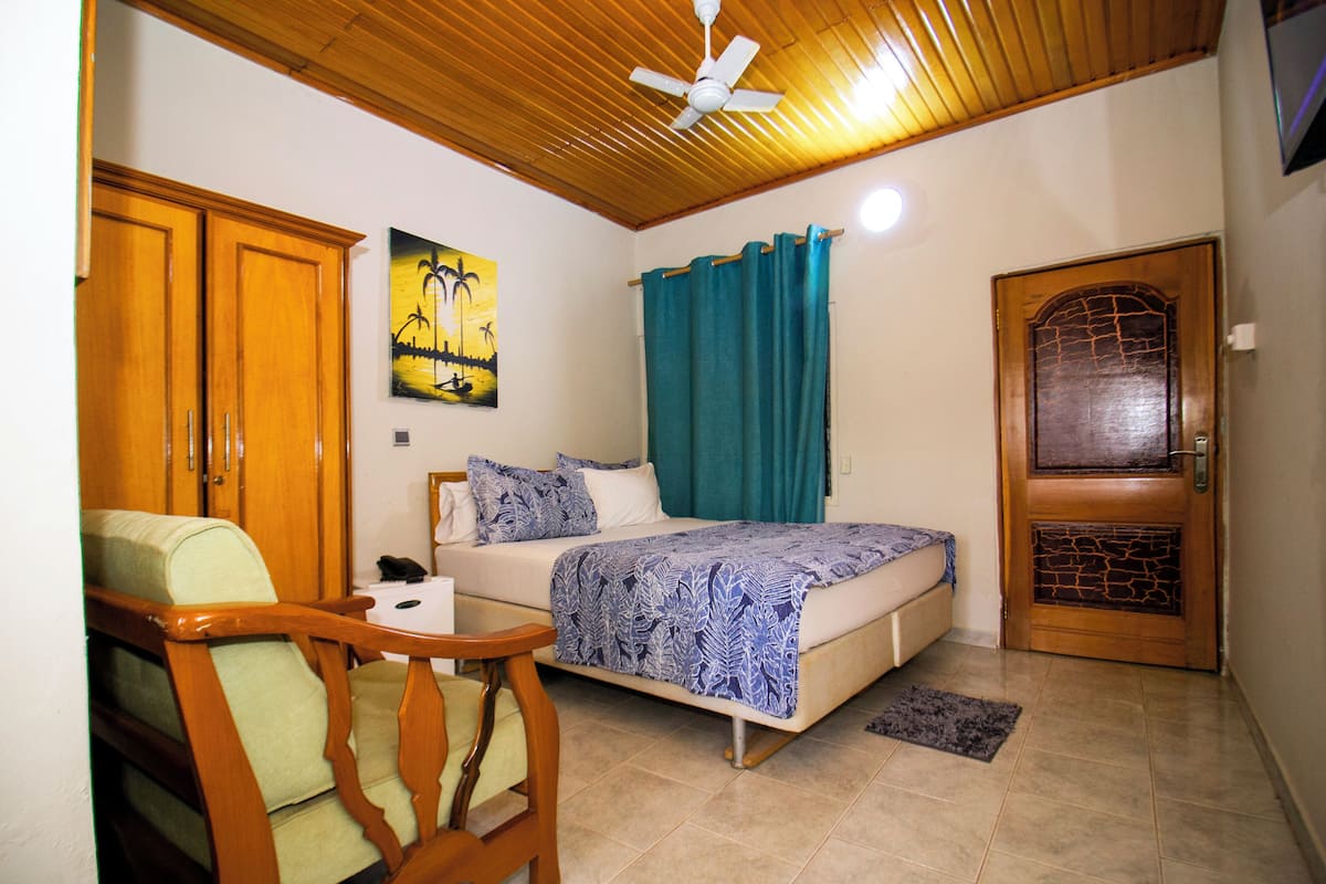 Quarto com cama, cadeira, escrivaninha, ventilador de teto, um quadro e porta com detalhe em pedra.