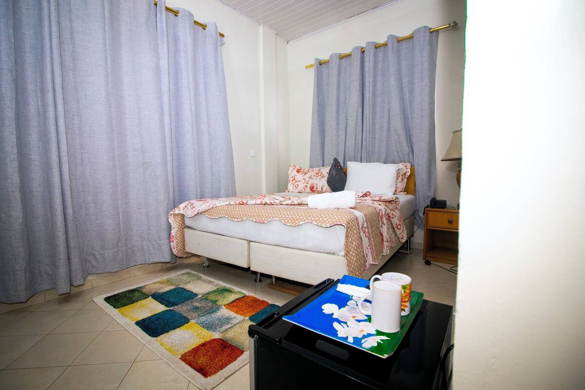 Quarto com cama, cortinas, mesa de cabeceira, abajur e tapete colorido.