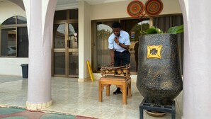 Un hombre ajusta una estructura de madera cerca de una gran maceta decorativa en un edificio con suelo de baldosas y columnas arqueadas.
