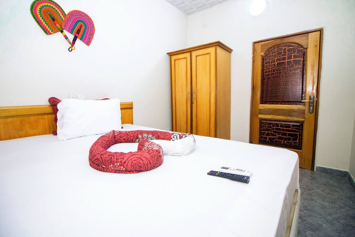 Cama bem arrumada com almofadas, um controle remoto e um guarda-roupa de madeira num quarto com porta e decoração na parede.