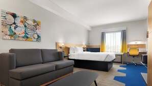 A hotel room with a gray sofa, a bed, a desk with a chair, and a large abstract painting on the wall.