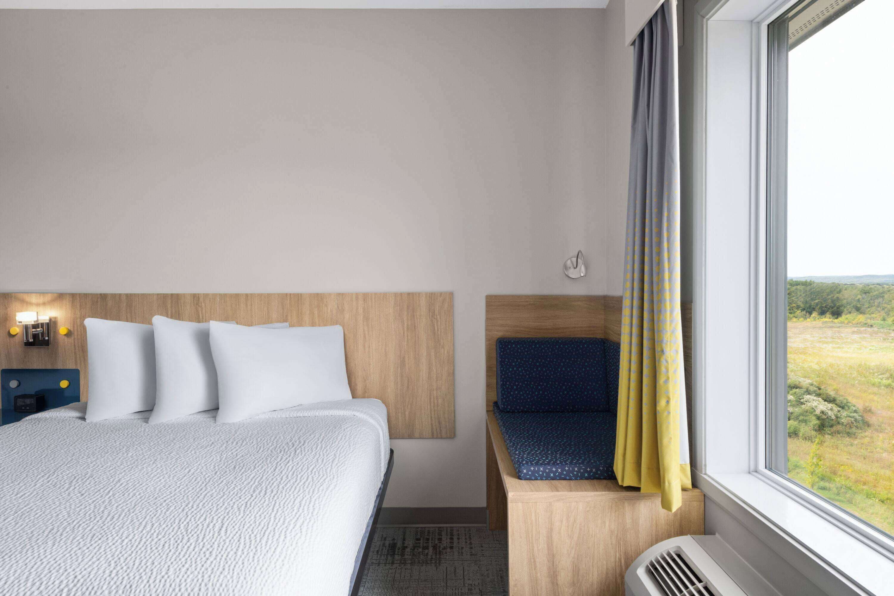 A hotel room with a bed, a bedside table with a lamp, a window with curtains, and a view of the outdoors.