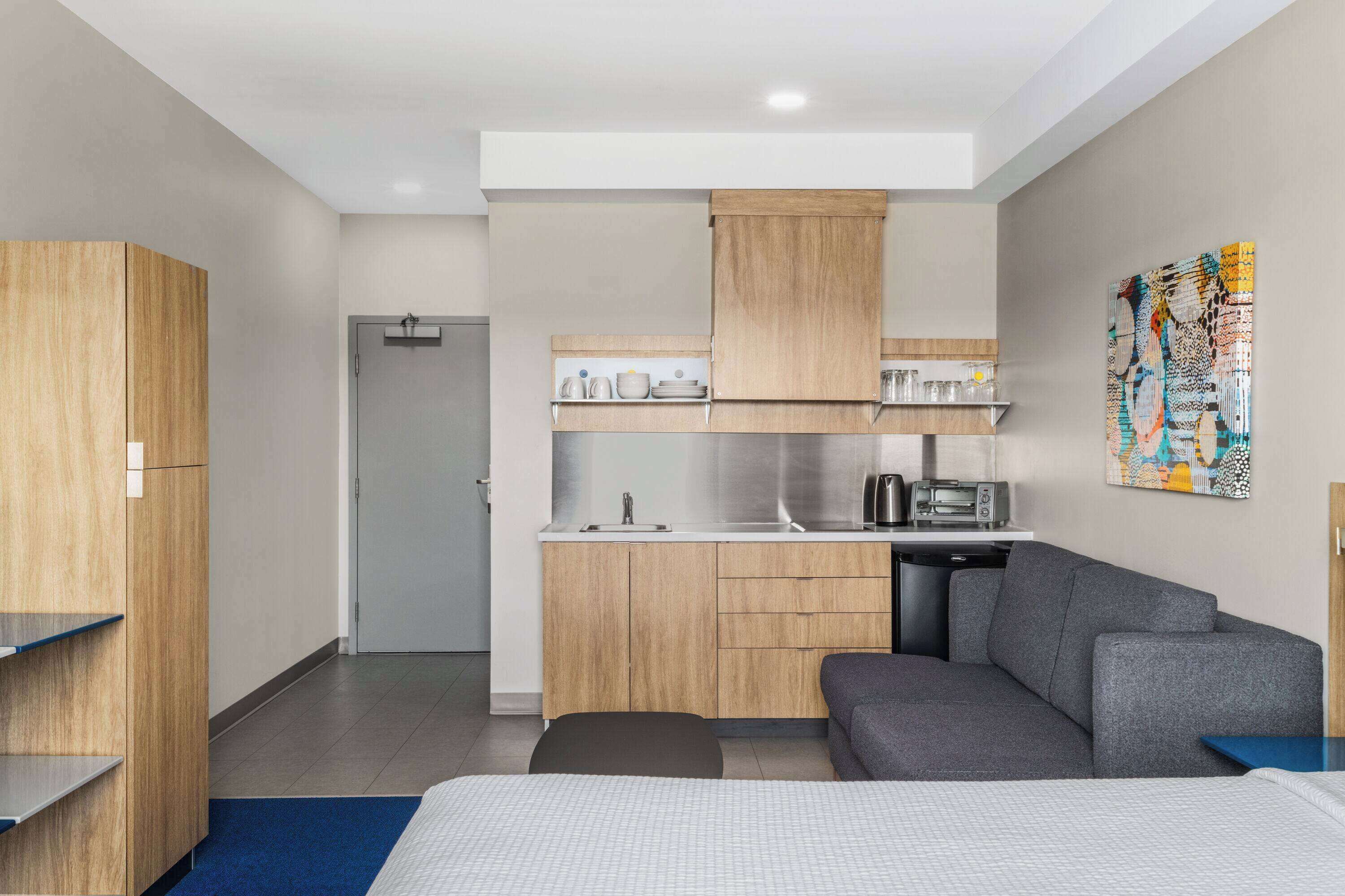 A modern hotel room with a kitchenette, a sofa, a bed, and a large abstract painting on the wall.