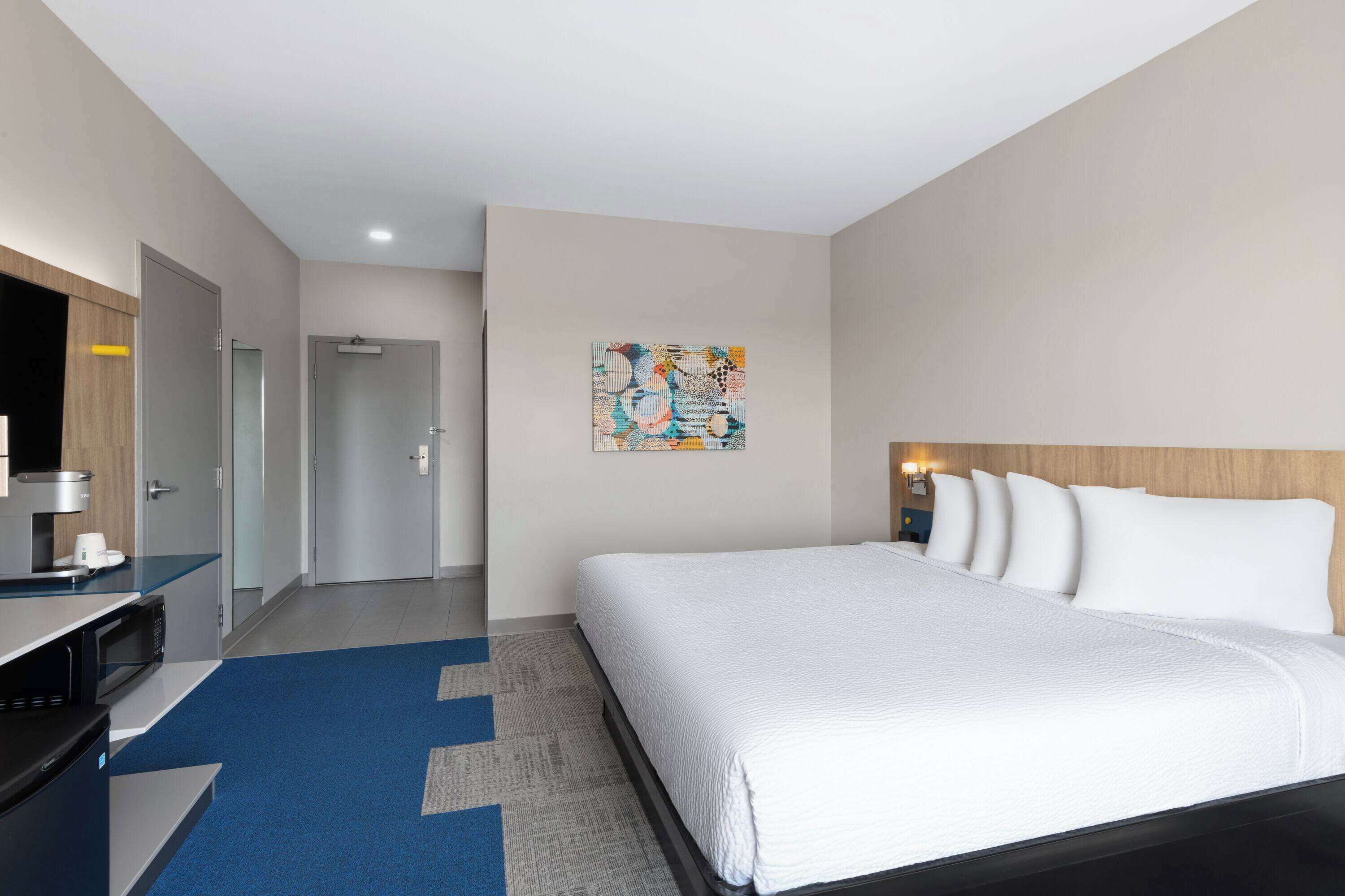 A hotel room with a bed, a desk with a computer, a microwave, a door, and a painting on the wall.