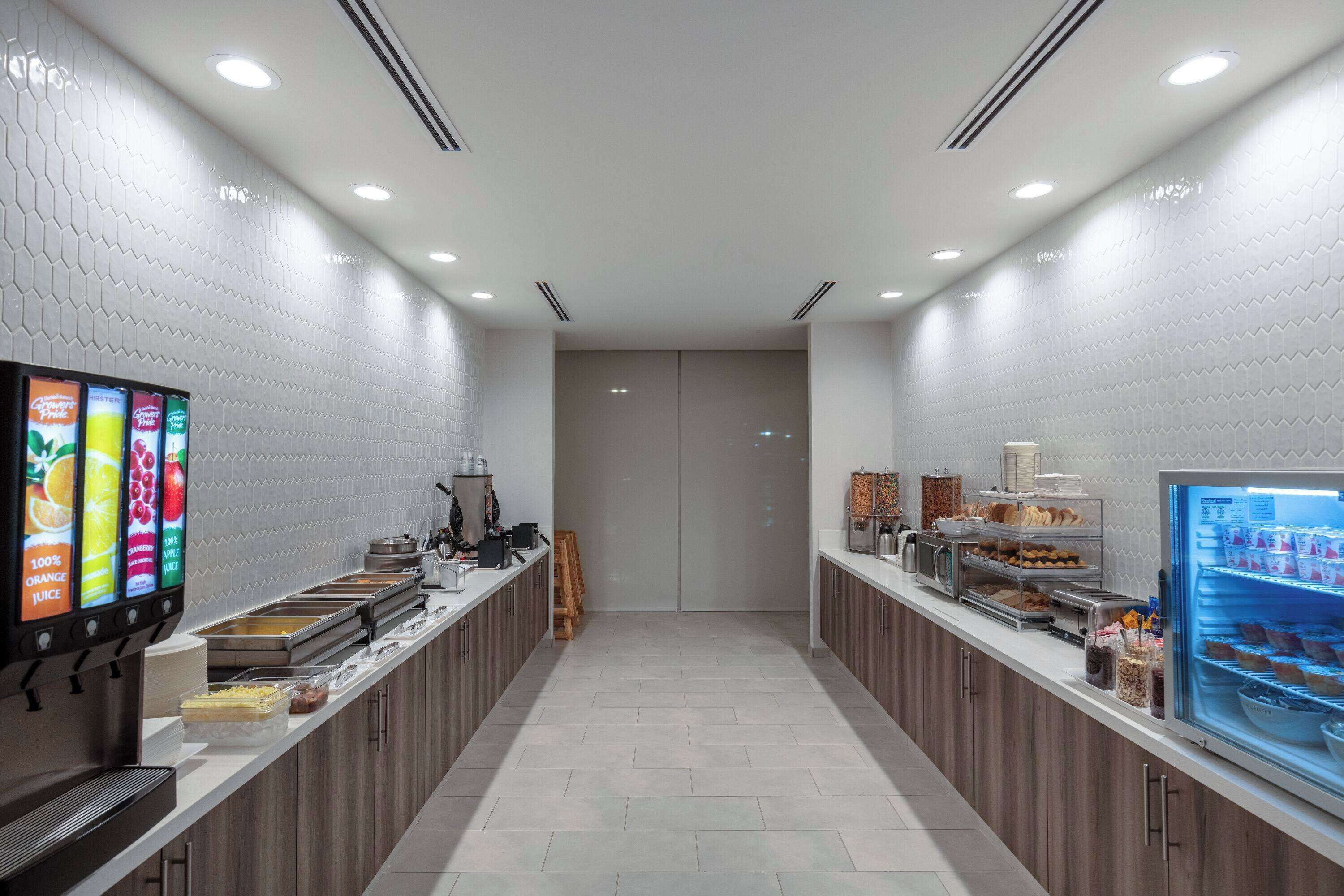 A modern, well-lit breakfast area with a self-serve beverage station, a refrigerated display case with pastries, and a clean, tiled floor.
