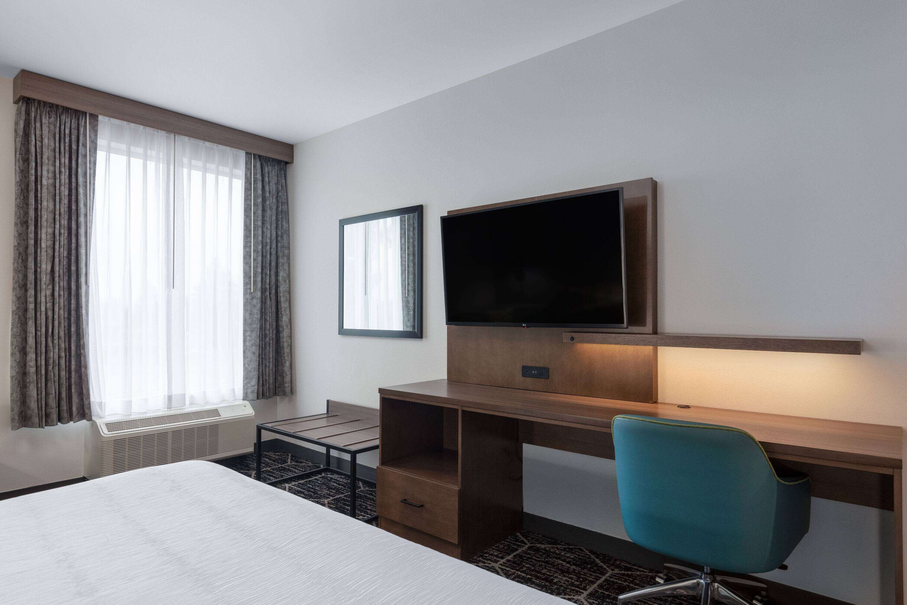 A hotel room with a bed, a desk with a chair, a TV, and a window with curtains.
