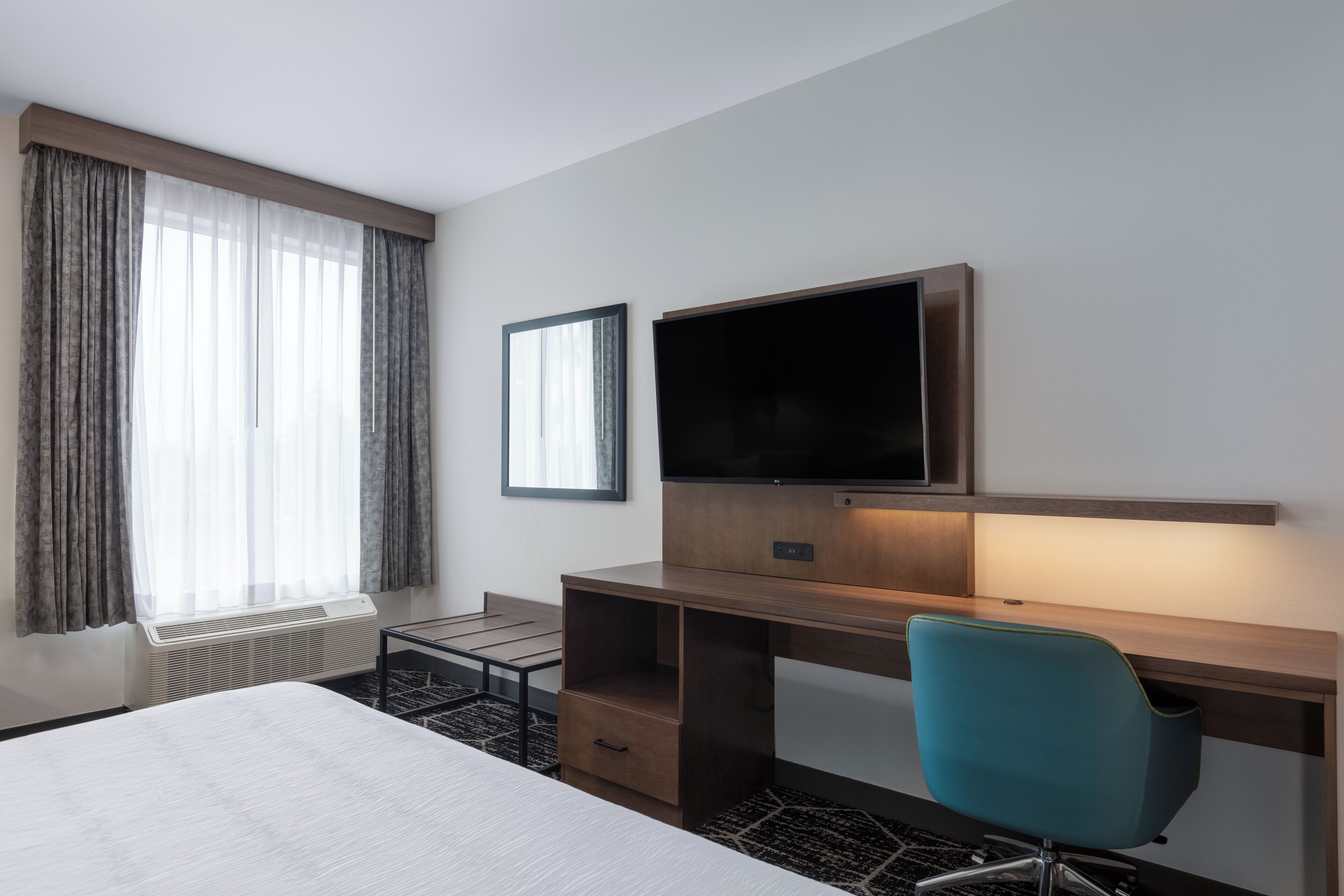 A hotel room with a bed, a desk with a chair, a flat-screen TV, and a window with curtains.