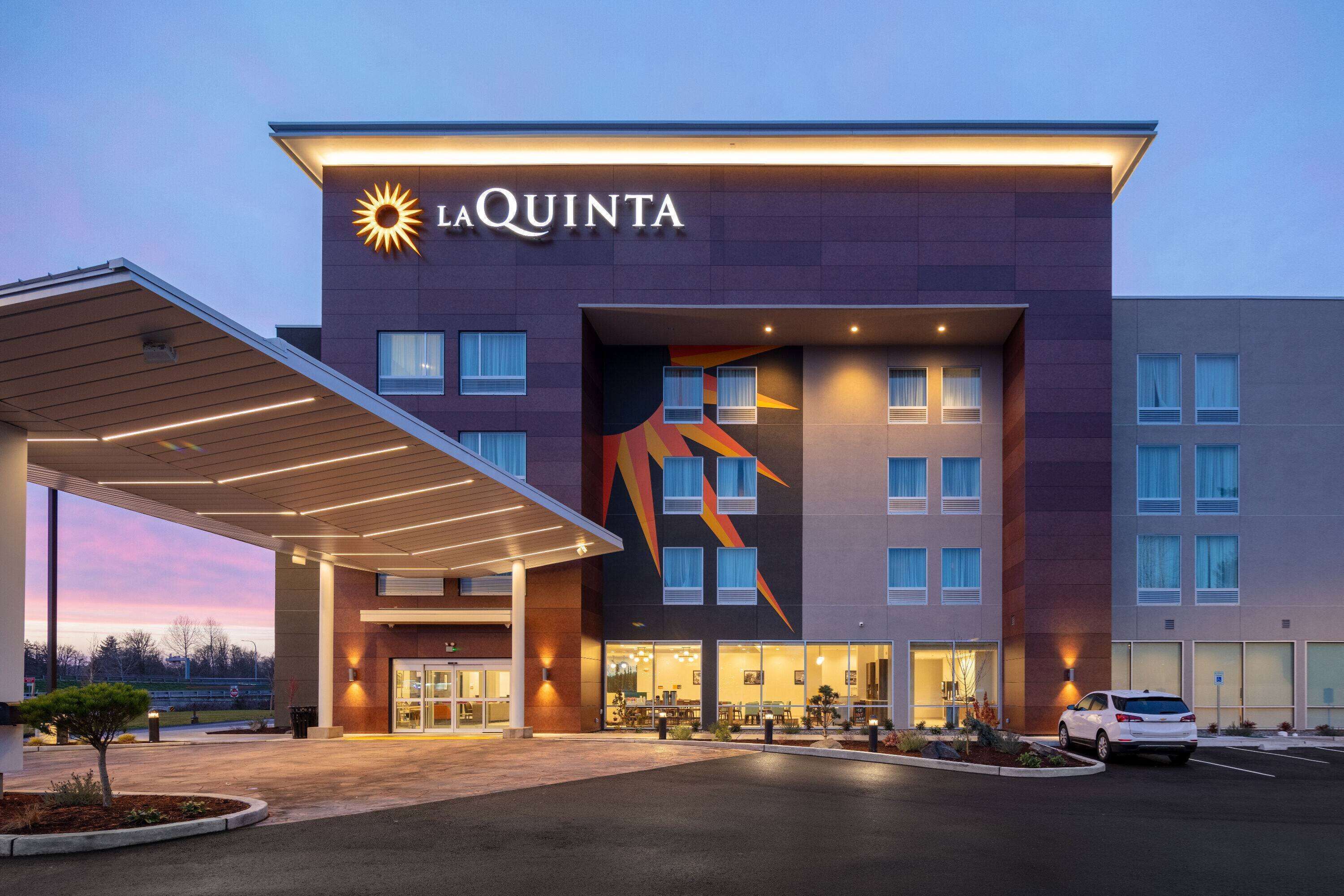 Un hôtel La Quinta au design moderne, avec un logo solaire bien visible, une entrée abritée et un espace de stationnement.