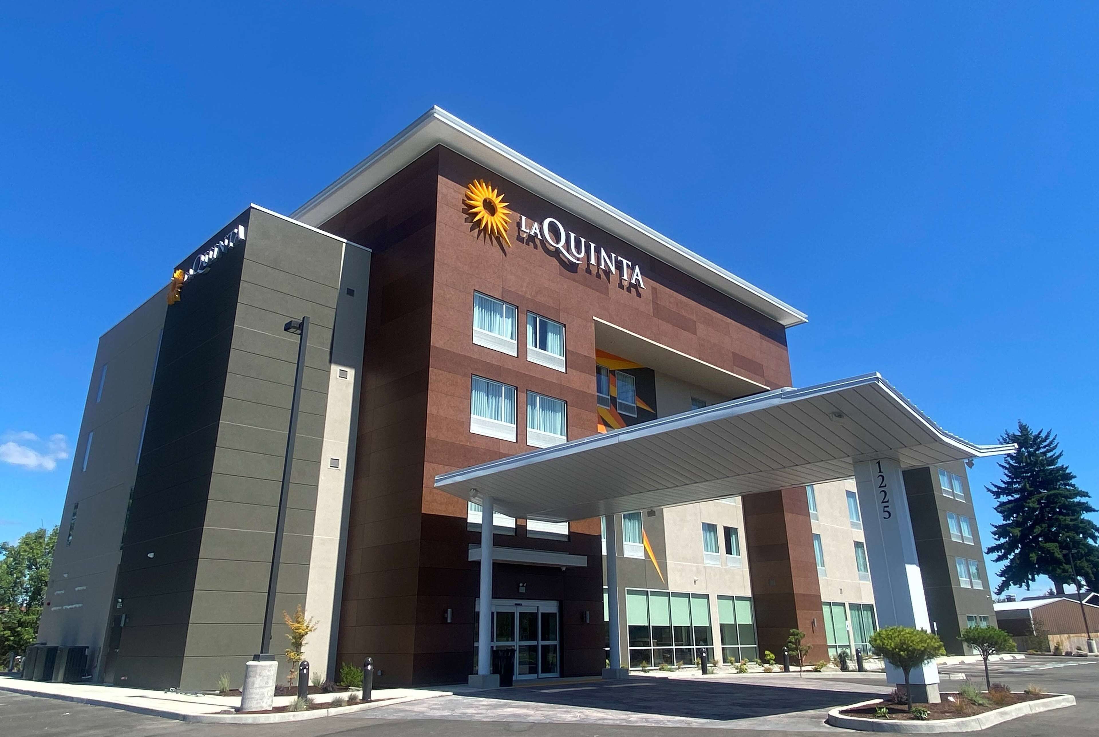 A La Quinta hotel with a modern design, featuring a prominent sun logo, a covered entrance, and a parking area with landscaping.