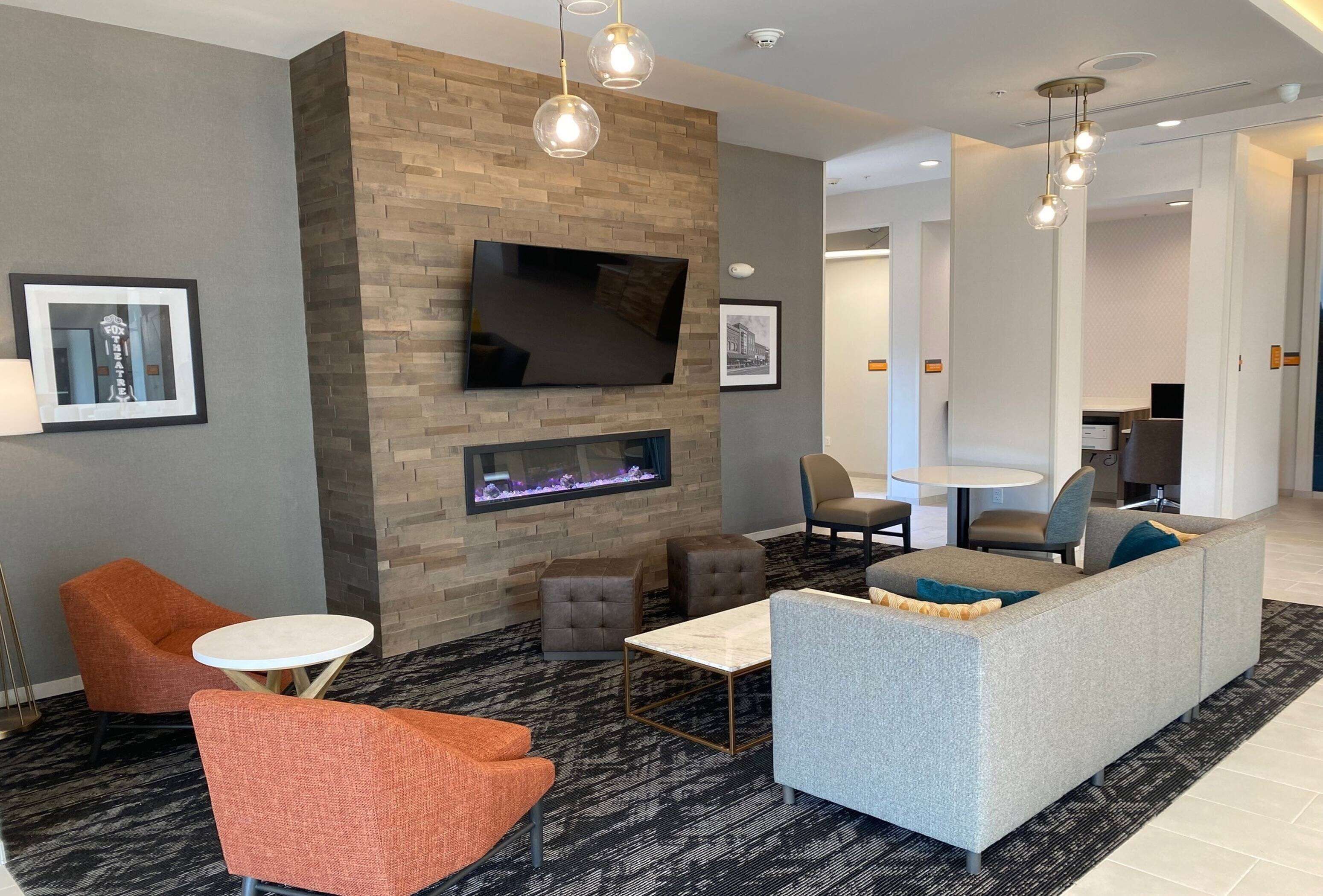 A modern living room with a flat-screen TV mounted on a brick wall, a fireplace, and contemporary furniture including a grey sofa and orange armchairs.