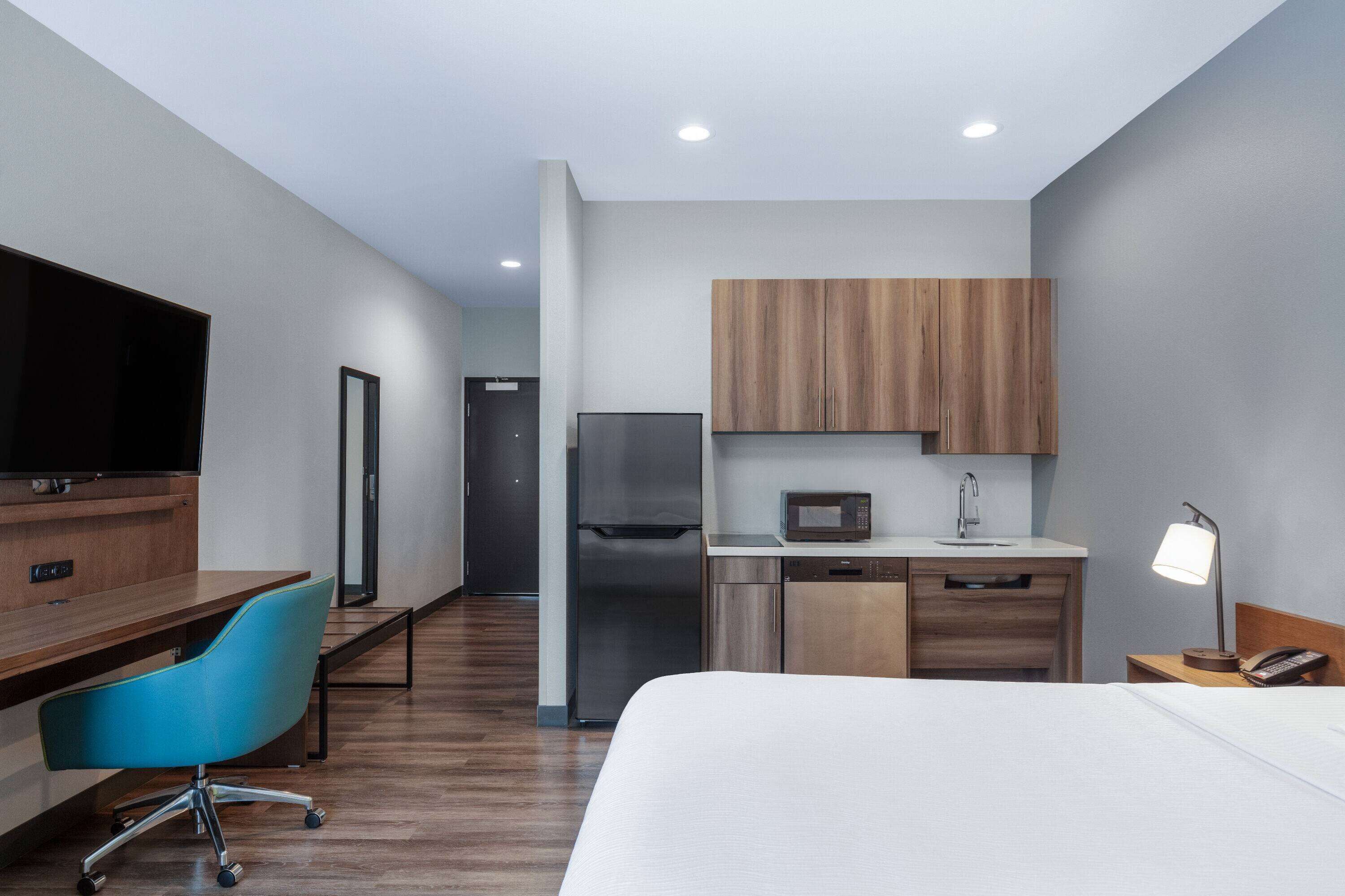 A modern hotel room with a bed, a desk with a chair, a kitchenette with a microwave, refrigerator, and sink, and a television.